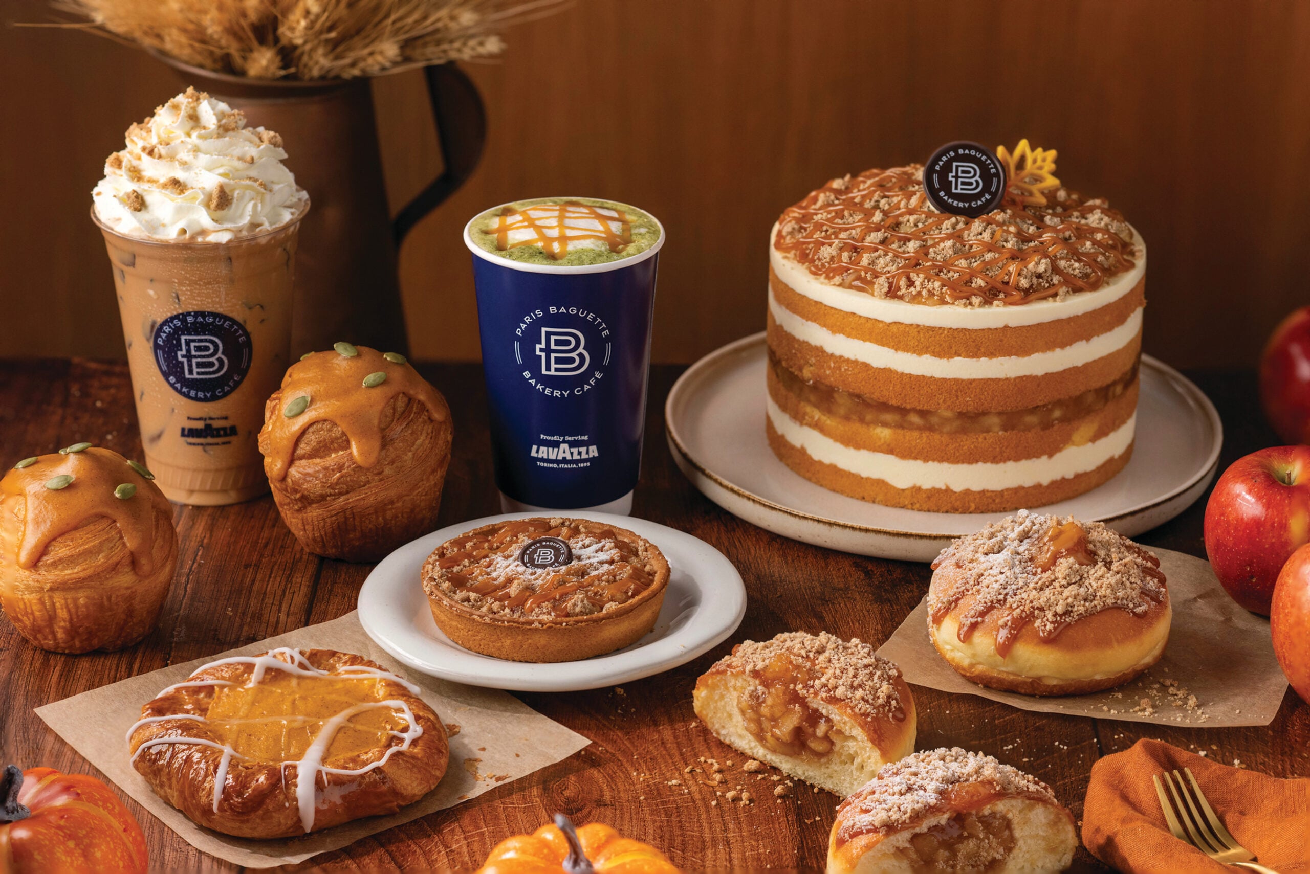 A variety of autumn-themed pastries, apple desserts, and two iced drinks are displayed on a wooden table with apples and a layered cake in the background.