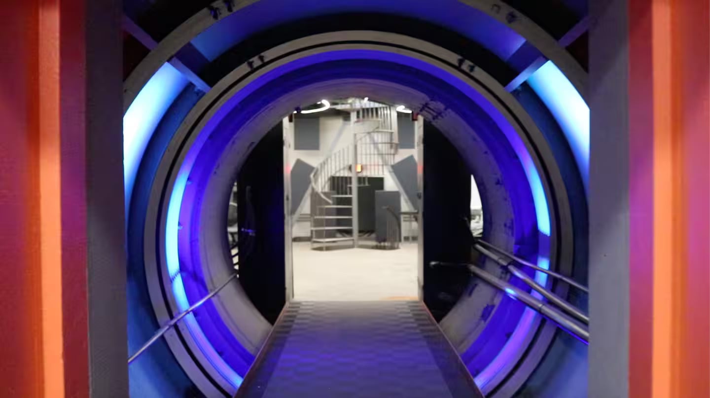 A futuristic circular doorway with glowing blue lights leads to a modern room with a spiral staircase in the background.