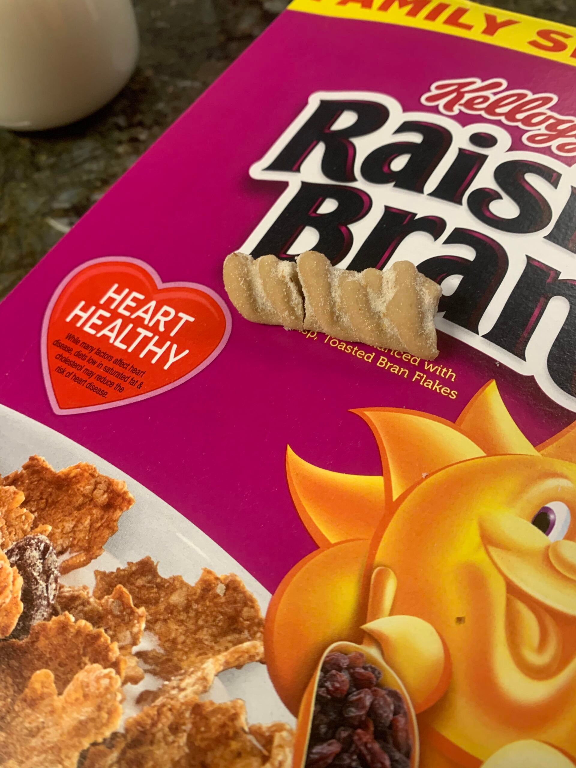 A piece of bran cereal is resting on top of a Raisin Bran cereal box near a red heart that says “Heart Healthy.” Part of a bowl of cereal and raisins is visible on the box.