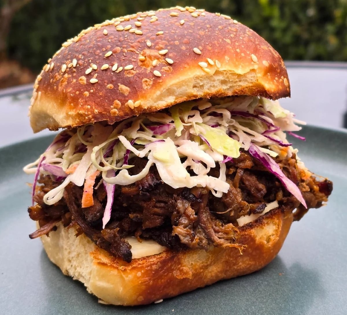 A close-up of a pulled pork sandwich topped with coleslaw on a sesame-seed bun, served on a gray plate with a blurred outdoor background.