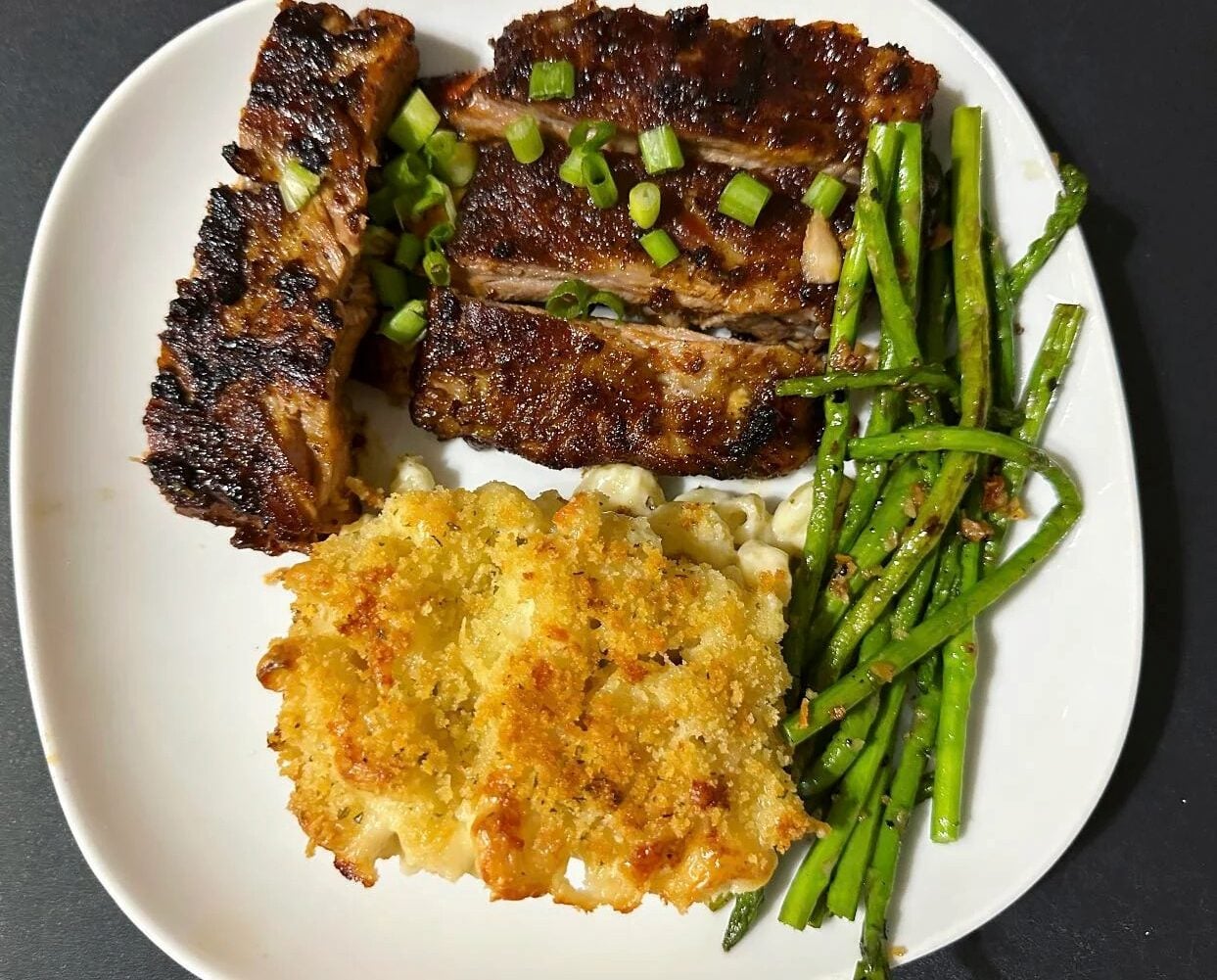 A white plate with three grilled ribs topped with chopped green onions, roasted asparagus spears, and a serving of crispy baked macaroni and cheese.