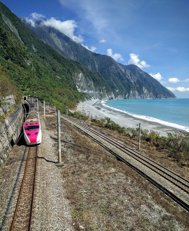 A pink and white train emerges from a tunnel beside a scenic coastline, with blue ocean waves and steep, green mountains under a bright, sunny sky.