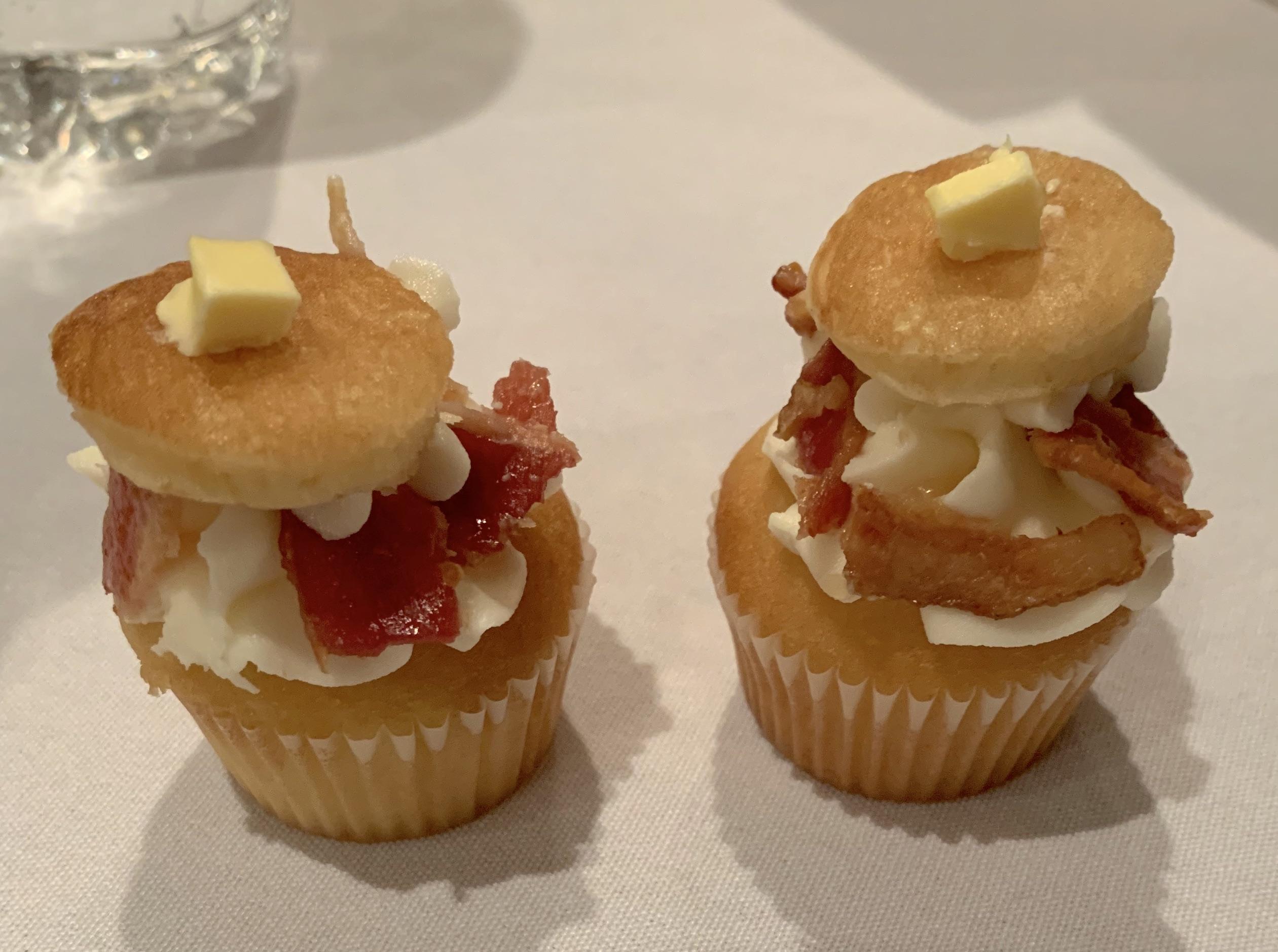 Two mini cupcakes topped with white frosting, crispy bacon pieces, a small biscuit, and a square of butter, sitting on a white tablecloth. A glass of water is visible in the background.