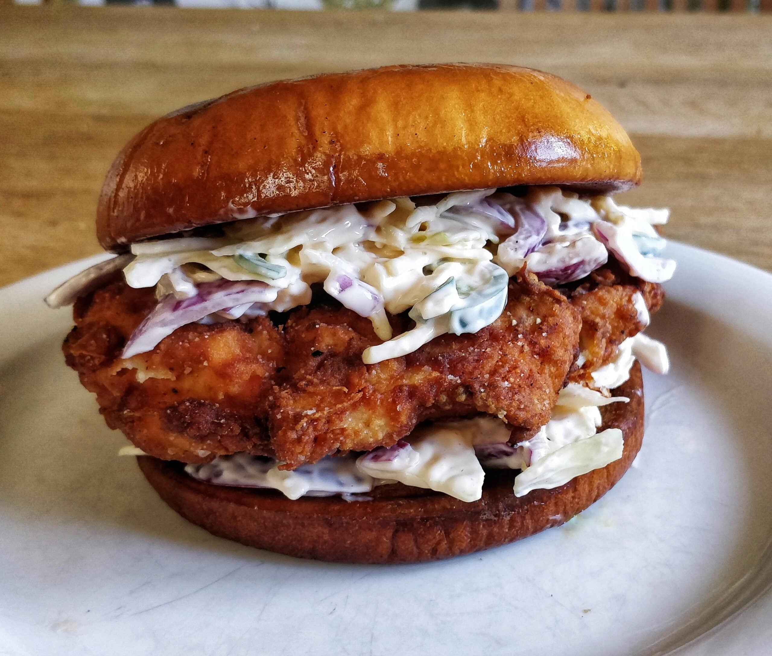 A crispy fried chicken sandwich with creamy coleslaw on a toasted bun sits on a white plate with a wooden table background.