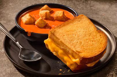 Grilled cheese sandwich on a black plate with a bowl of tomato soup topped with croutons. A spoon rests beside the bowl.
