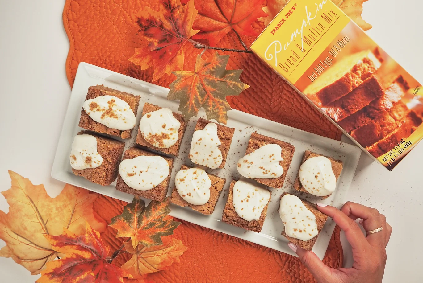 A hand reaches for a piece of pumpkin bread topped with whipped cream on a tray. The tray sits on an orange leaf-patterned mat with decorative autumn leaves and a box of pumpkin bread mix nearby.