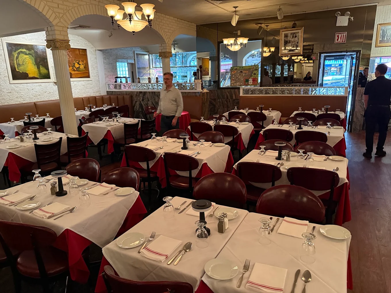 A cozy restaurant dining room with tables covered in white tablecloths, set with plates, glasses, and cutlery. Two people stand inside, and artwork decorates the walls. A large mirror reflects the scene.