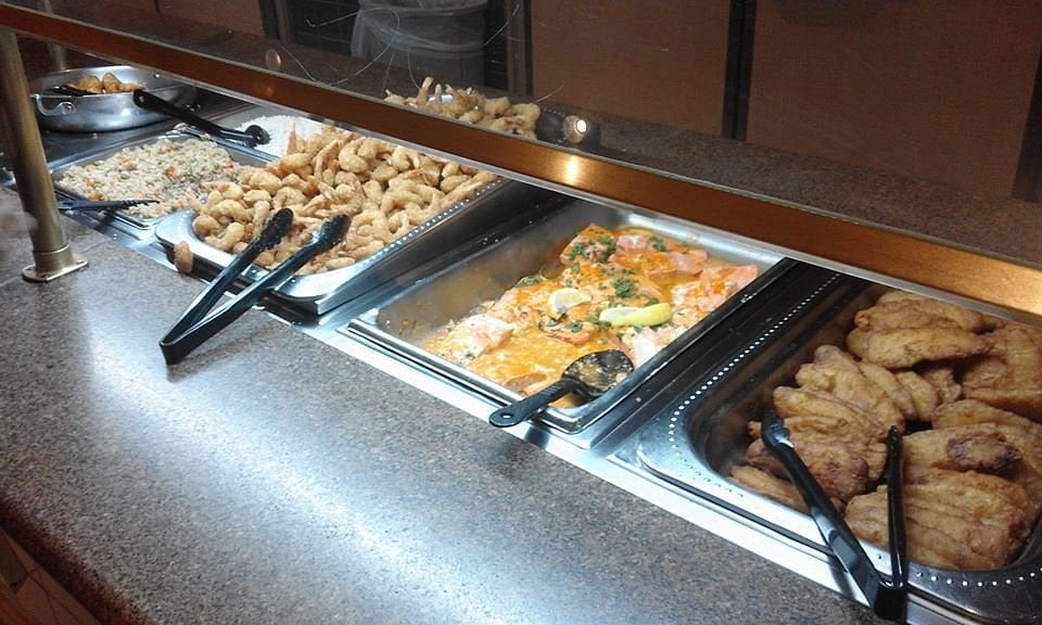 A buffet counter displays fried rice, breaded shrimp, salmon with lemon and herbs, and fried fish fillets, each in metal trays with serving tongs on a granite countertop.