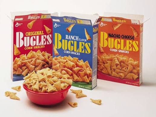Three boxes of Bugles corn snacks—Original, Ranch Flavor, and Nacho Cheese—are behind a red bowl filled with Bugles. Some Bugles pieces are scattered around the bowl on a white surface.