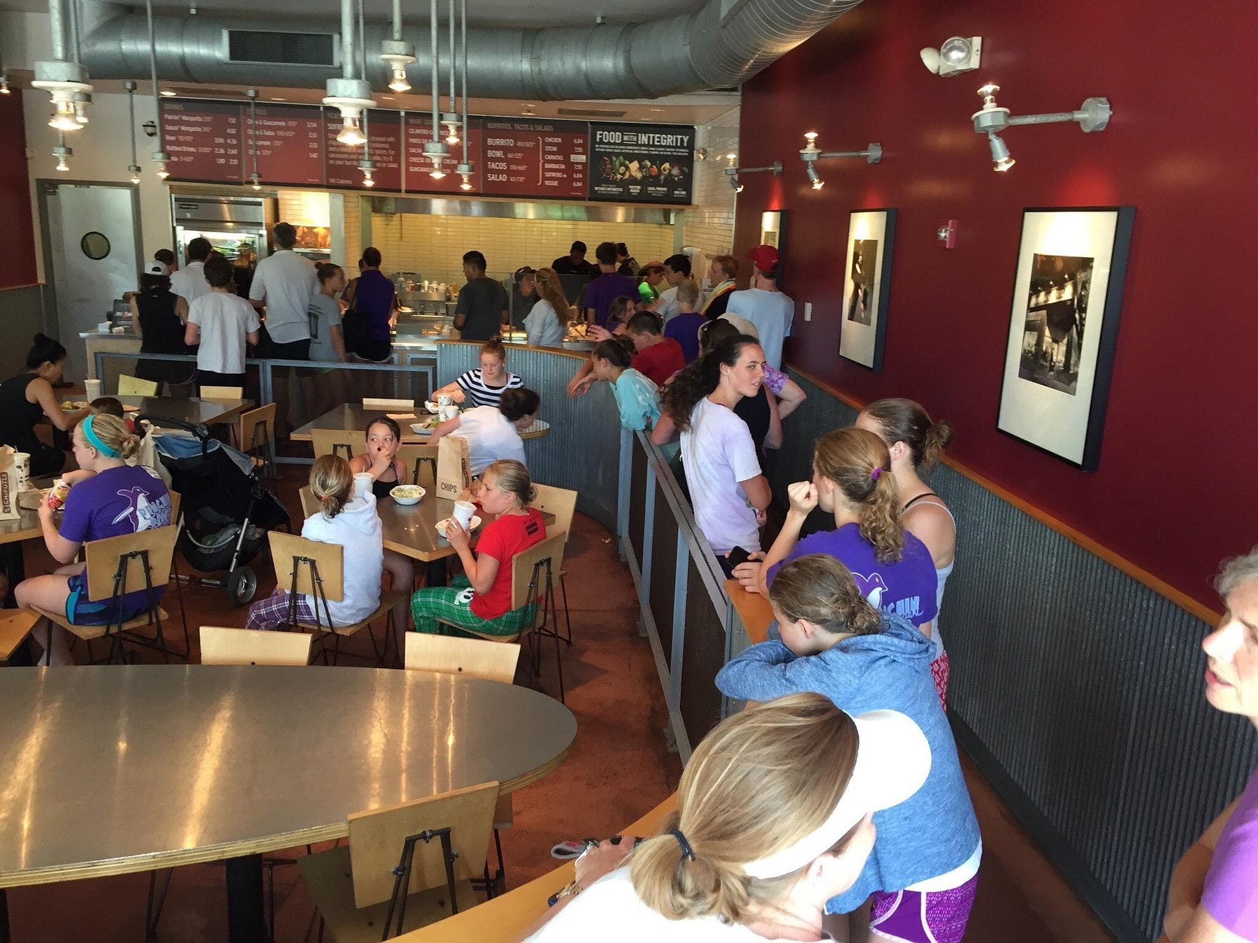 People in athletic clothes are lined up and seated inside a busy casual restaurant with metal and wood decor, eating, talking, and waiting to order food at the counter.