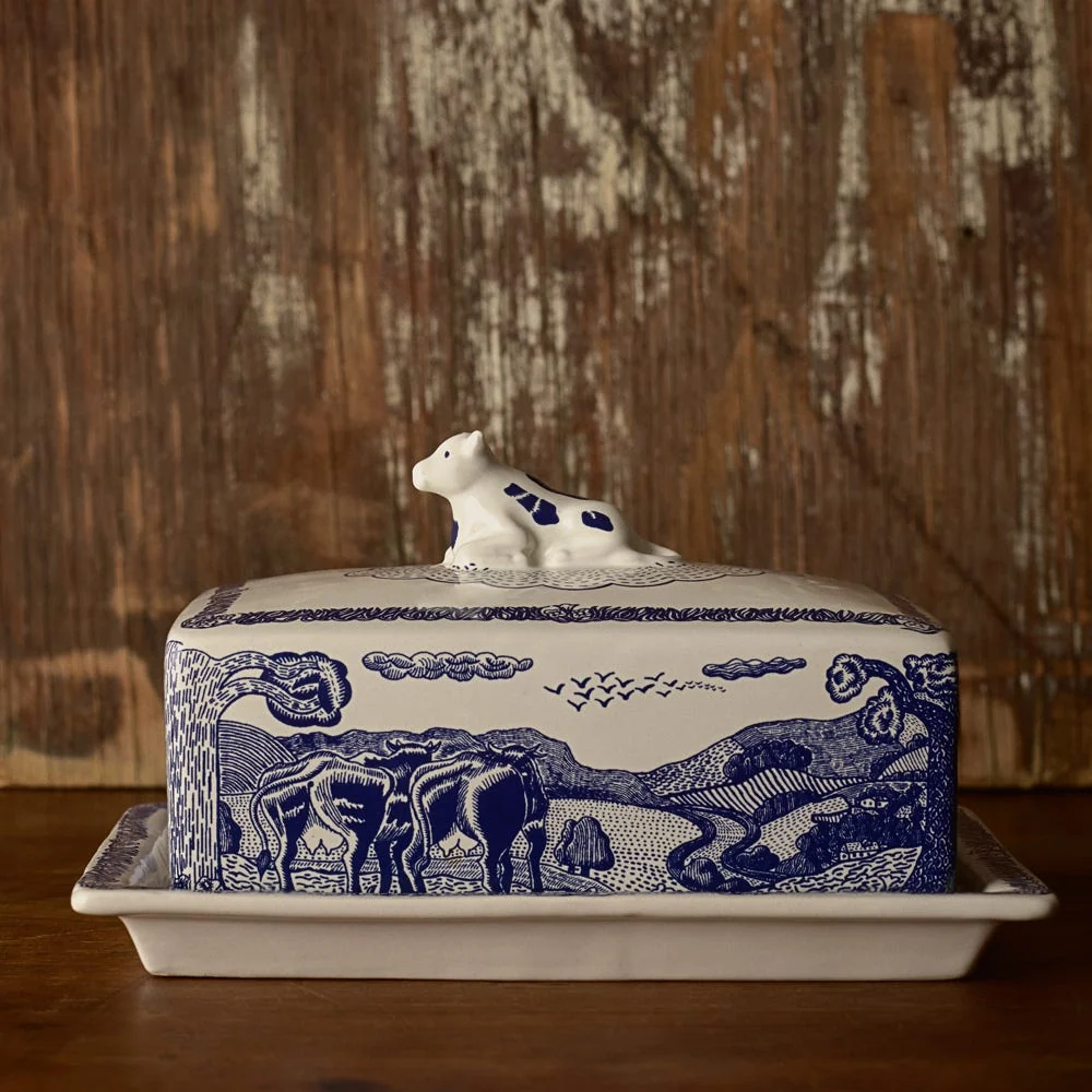 A rectangular ceramic butter dish with blue pastoral designs and a lid topped with a small cow figurine, set against a rustic wooden background.