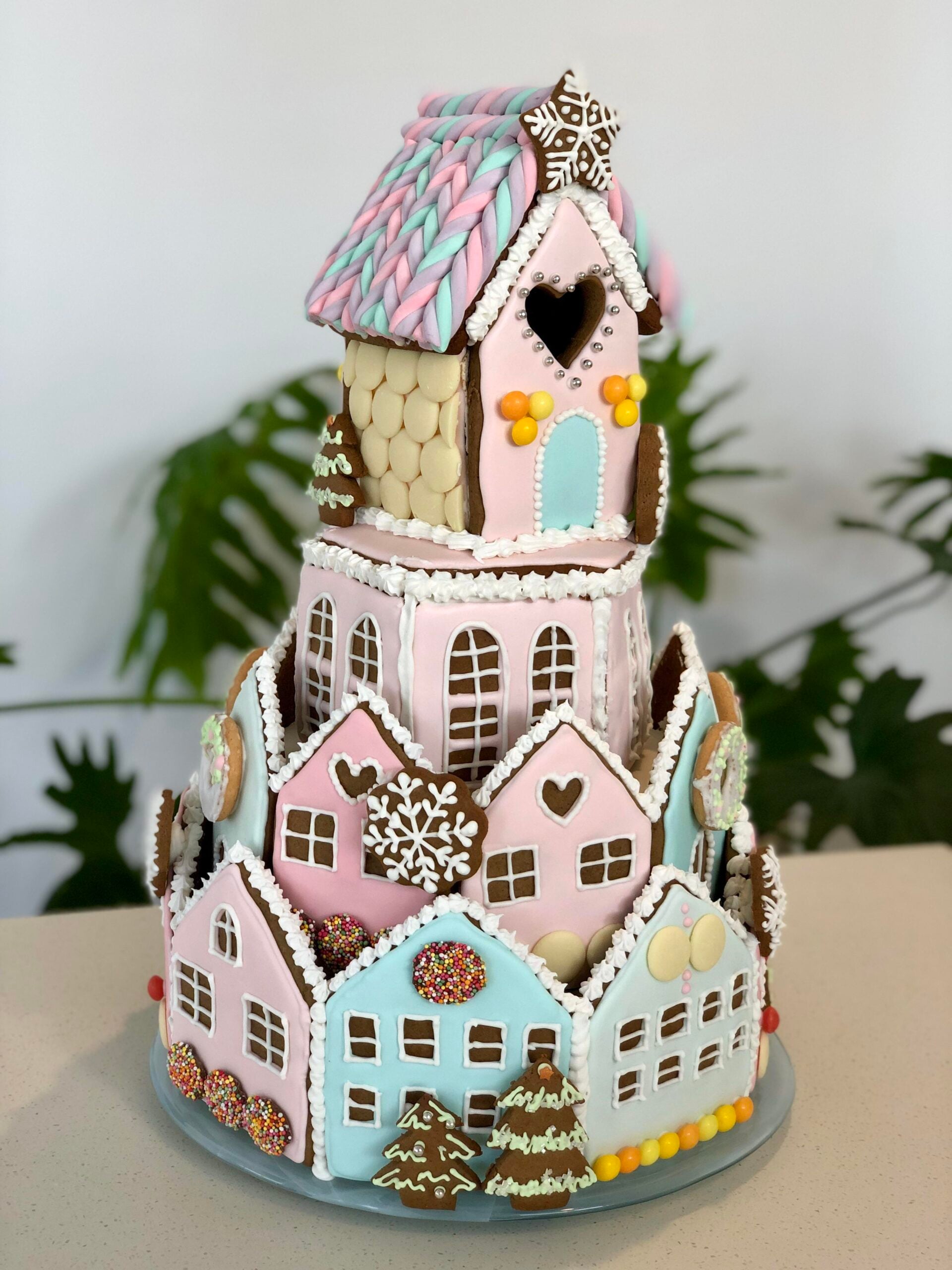 A colorful, multi-tiered gingerbread house decorated with pastel icing, candies, and cookies—reminiscent of homemade Christmas treats—features heart and snowflake designs as it sits on a plate with green plants blurred in the background.