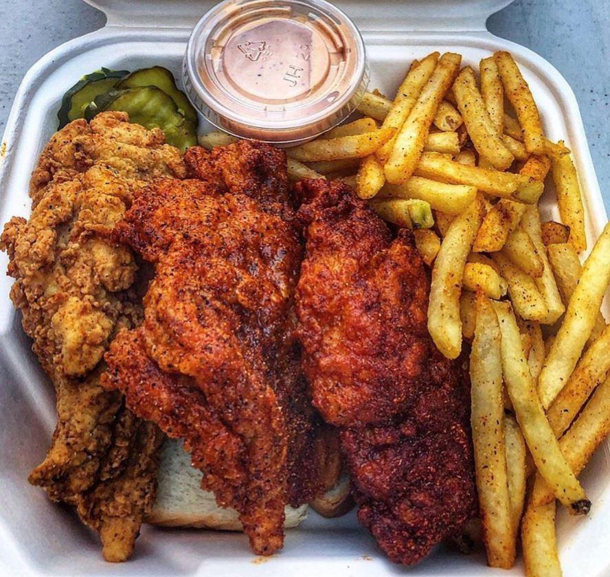 A takeout container with seasoned fries, fried chicken tenders—some plain, some spicy—a few pickles, a slice of white bread, and a small container of dipping sauce.