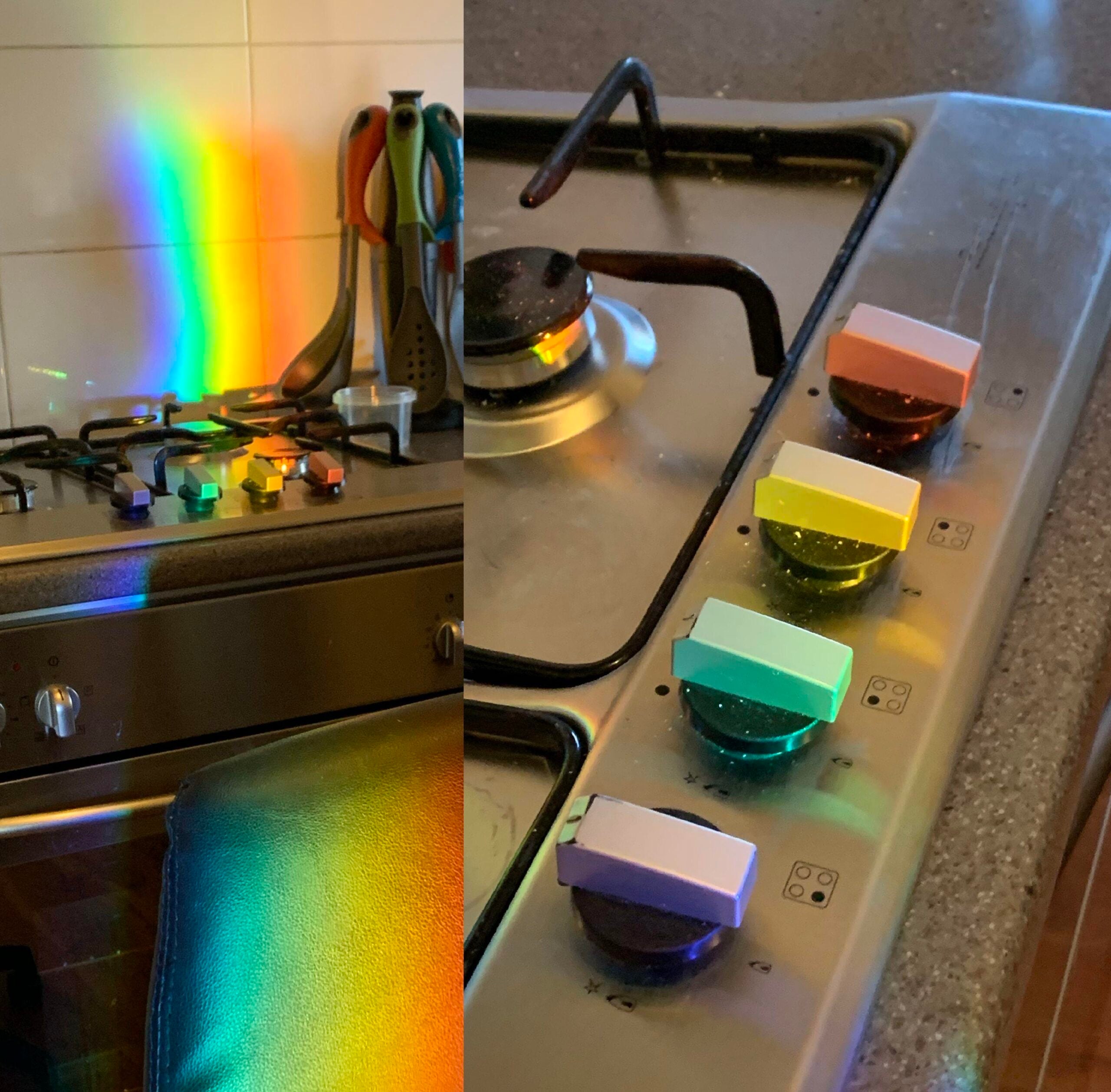 A kitchen stove with rainbow light reflections on knobs, countertop, and drawers. Four stove knobs each have a different colored reflection: pink, yellow, green, and blue. Utensils are seen in the background.