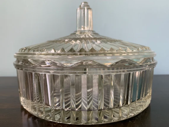 Clear glass candy dish with a lid, featuring vertical and zigzag patterns. The dish has a faceted knob on top and sits on a dark surface with a light blue background.