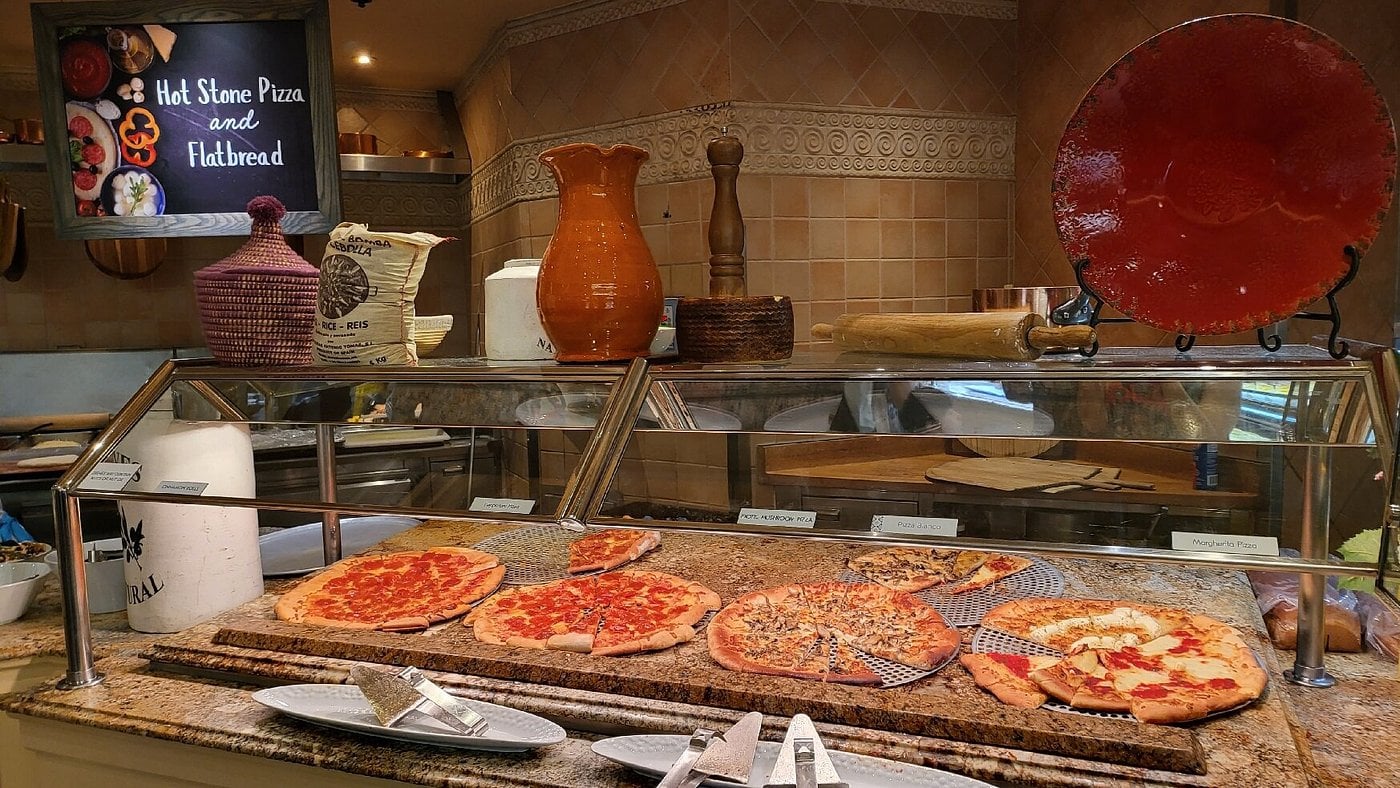 A buffet counter displays six types of pizza on trays behind glass. Plates, tongs, and serving utensils are in front. Behind the counter are ceramic jars, kitchen tools, and a sign reading “Hot Stone Pizza and Flatbread.”