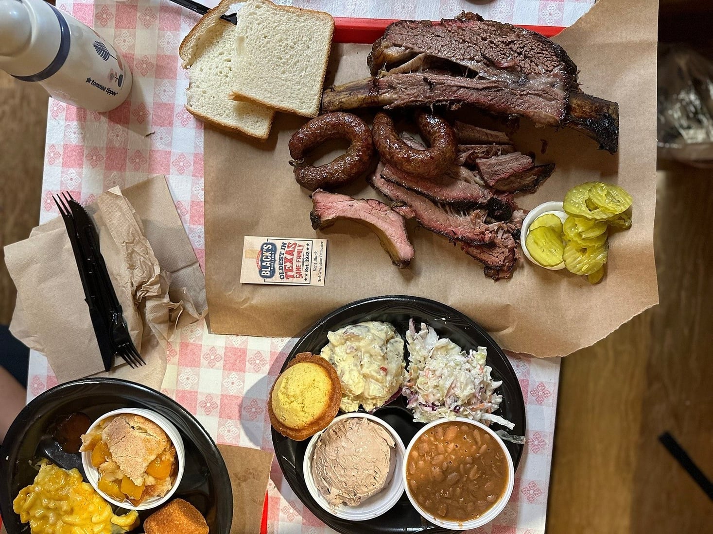 Barbecue meal on brown paper with brisket, sausage, pickles, white bread, and sides including cole slaw, baked beans, cornbread, and peach cobbler, on a checkered tablecloth. Plastic utensils and napkins are nearby.