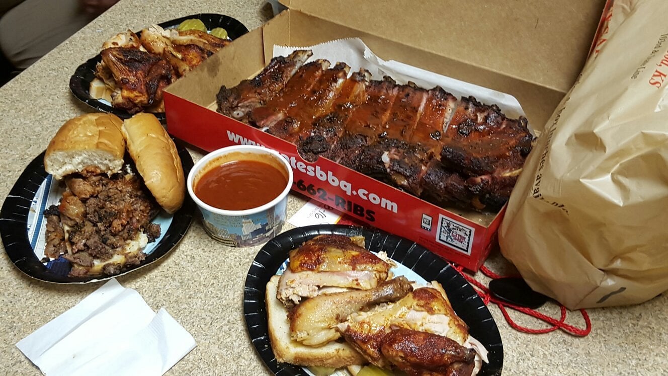Barbecue meal with a box of ribs, plates of roasted chicken, pulled pork with sandwich buns, and a cup of barbecue sauce on a countertop. A plastic bag and napkins are also visible.