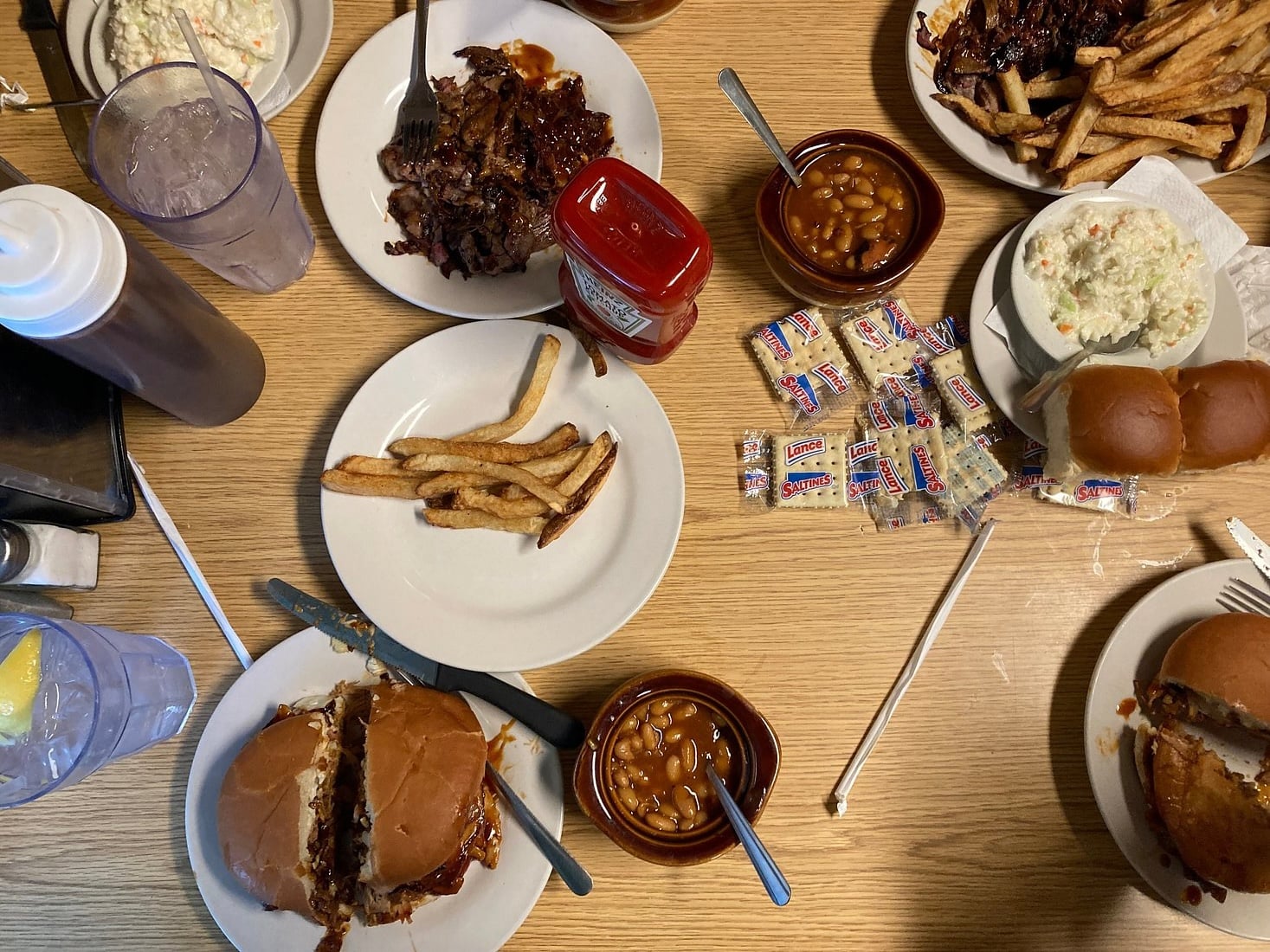 Top-down view of a table with barbecue plates, buns, fries, baked beans, coleslaw, water with lemon, ketchup, napkins, and packets of crackers on a light wooden surface.