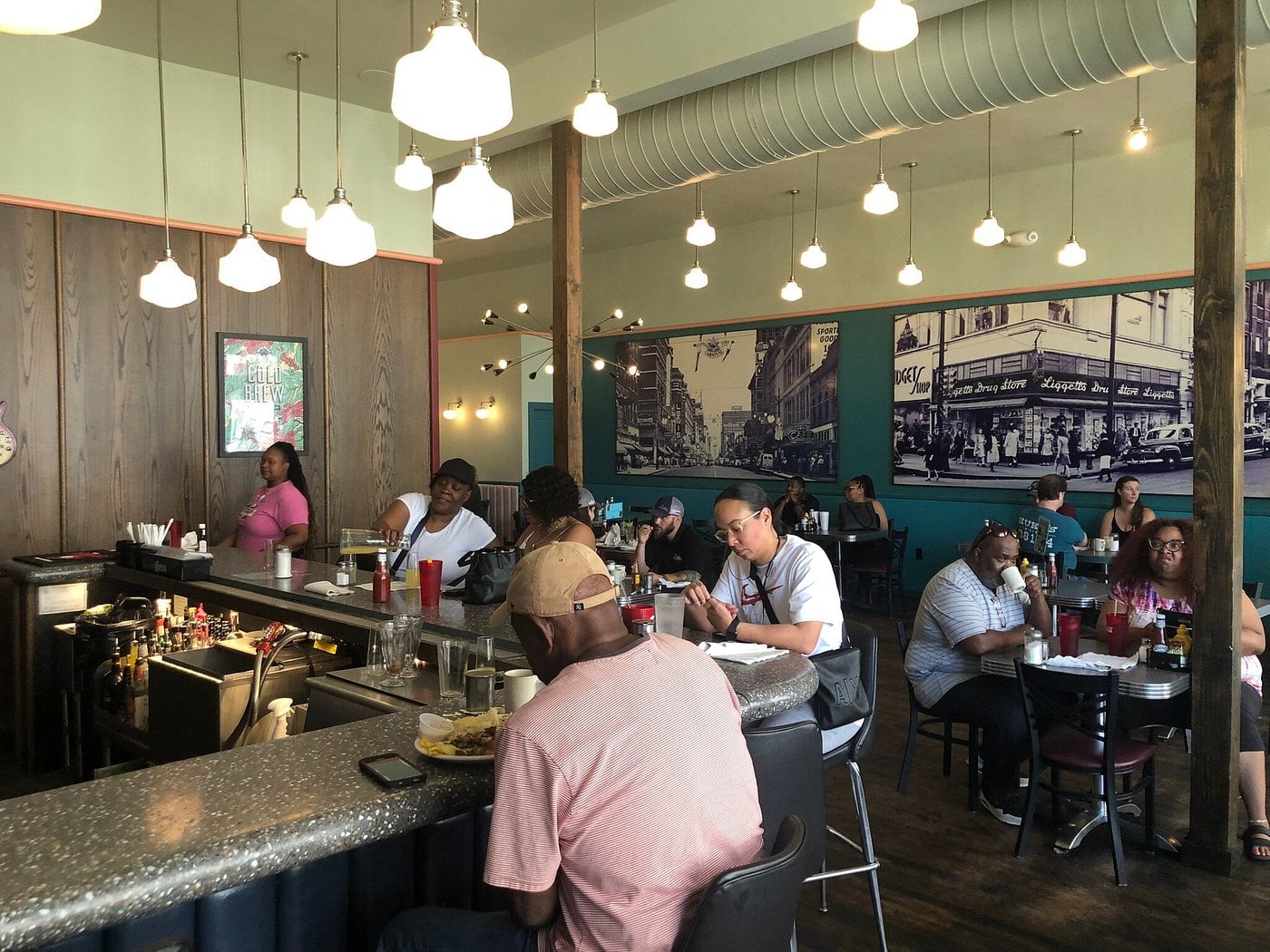 Diners sit at tables and a counter in a casual restaurant with hanging lights, wood accents, and large black-and-white city photos on the walls.