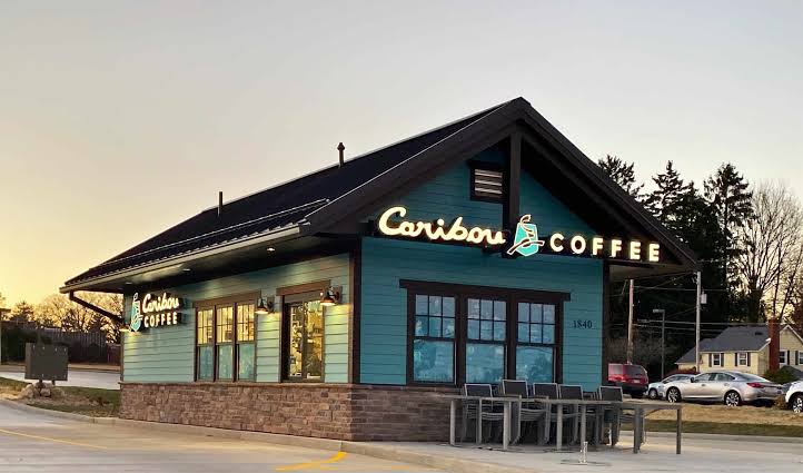 A teal and brown Caribou Coffee shop with outdoor seating and large illuminated signs, set against a sunset sky with trees and houses in the background.