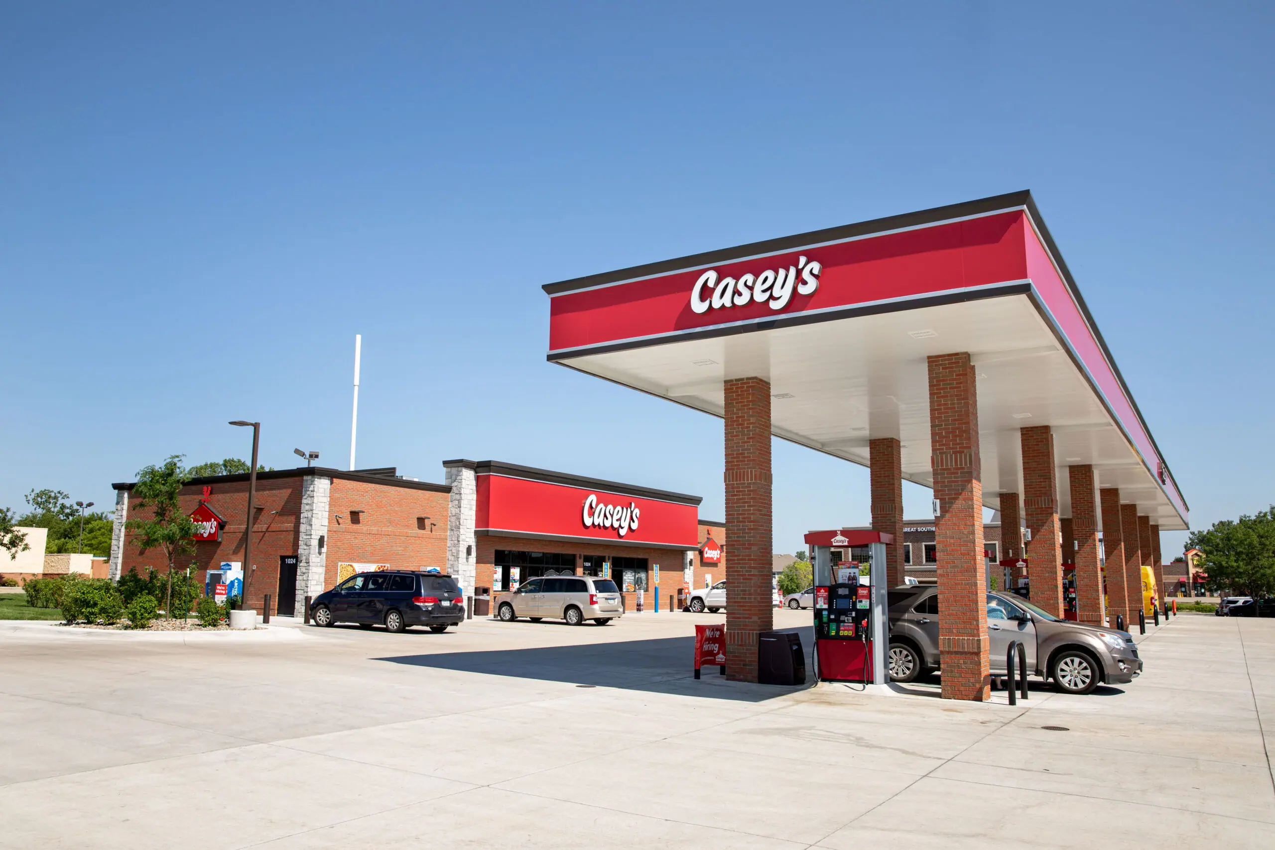 A Casey’s convenience store and gas station with several gas pumps, cars parked at the pumps, and clear blue sky overhead. The store and canopy feature red signs with white Casey’s lettering.
