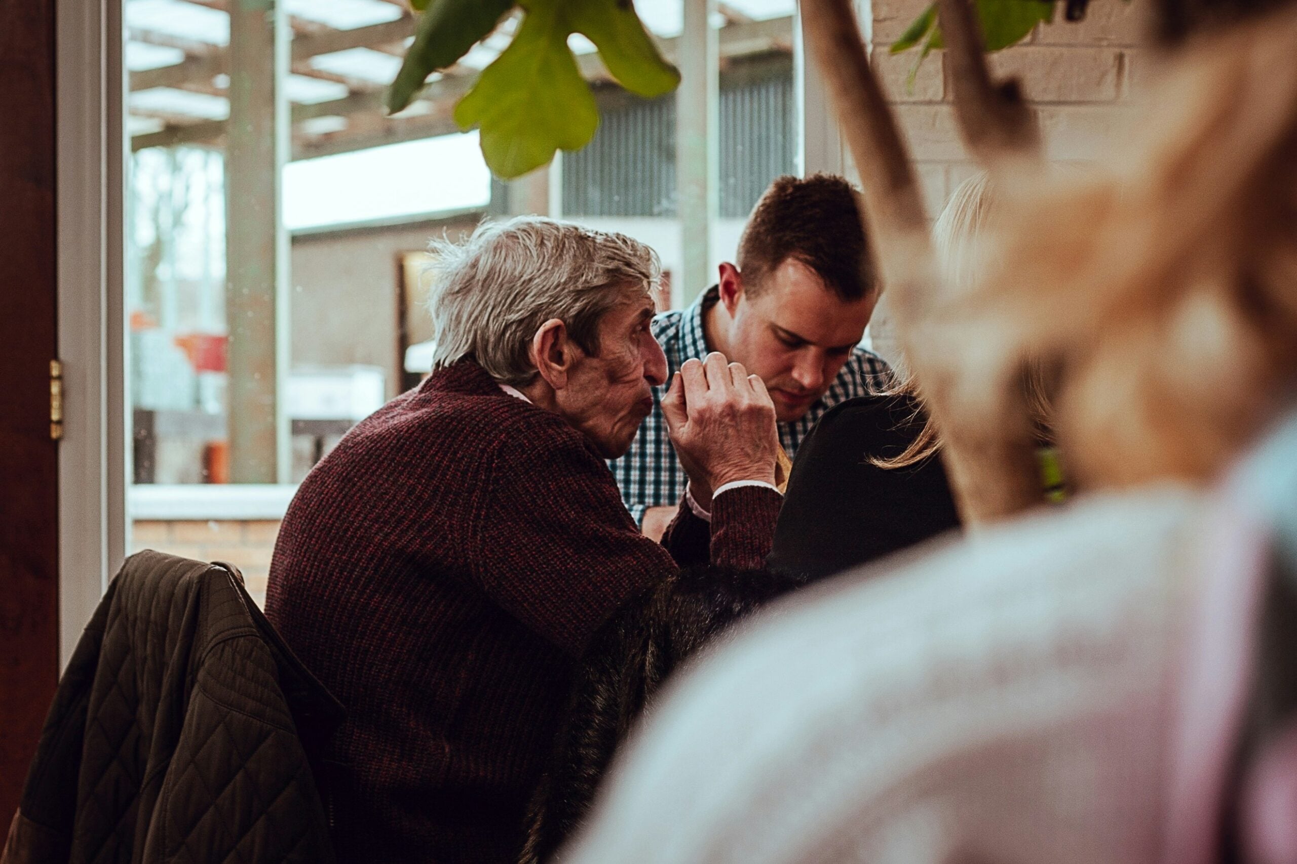 An elderly man and a younger man sit closely together at a table indoors, engaged in conversation about life in 2026. Blurred figures and greenery are visible in the foreground and background.