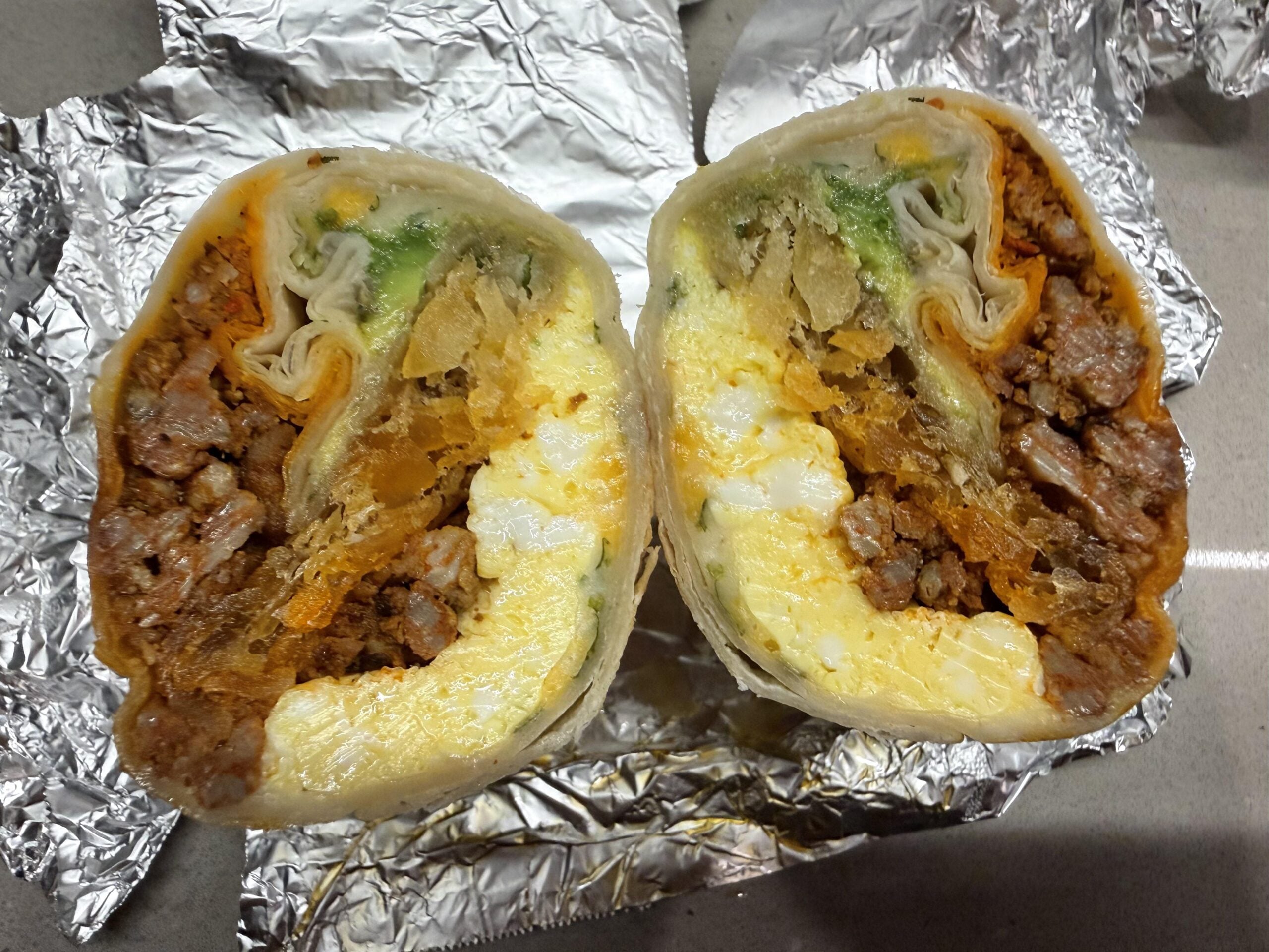 A breakfast burrito cut in half, showing layers of egg, beef, cheese, crispy potatoes, lettuce, and wrapped in a tortilla, sitting on aluminum foil.