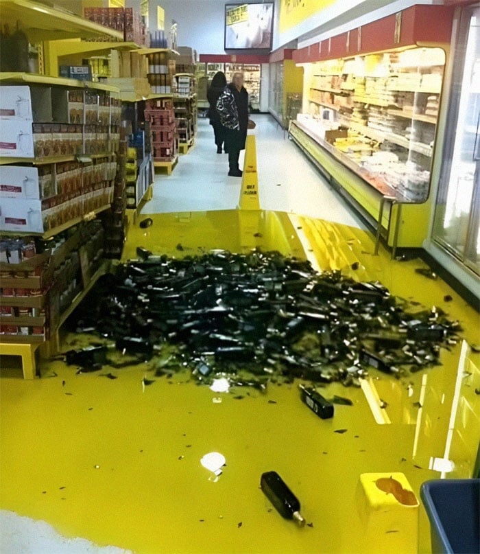 A large puddle of yellow liquid and many broken glass bottles cover a supermarket aisle floor. A caution sign is placed nearby, and a few people stand in the background.