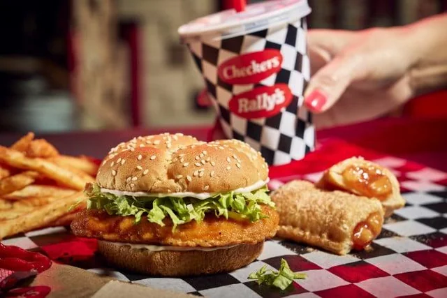 A Checkers & Rally’s meal with a crispy chicken sandwich, fries, two apple pies, and a checkered cup with a red straw. A hand reaches for the drink. The food is served on a red and black checkered tray liner.