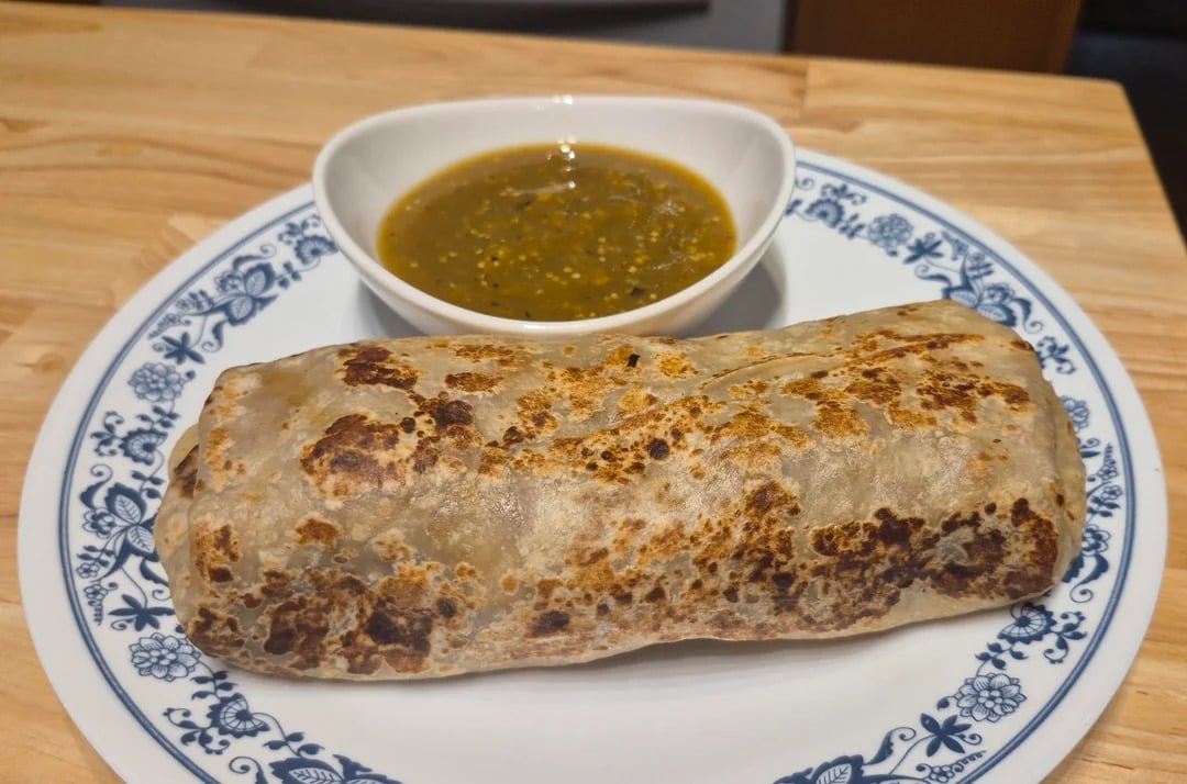 A grilled burrito sits on a white plate with blue designs, served alongside a small bowl of green salsa on a wooden table.
