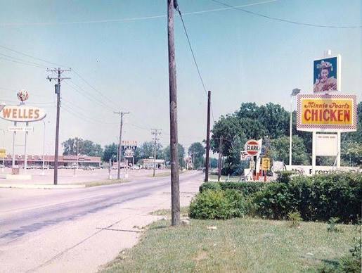 A vintage street scene with a “Mama Ford’s Chicken” sign, a Clark gas station, power lines, and other businesses, including a Welles sign, on a sunny day with clear skies.