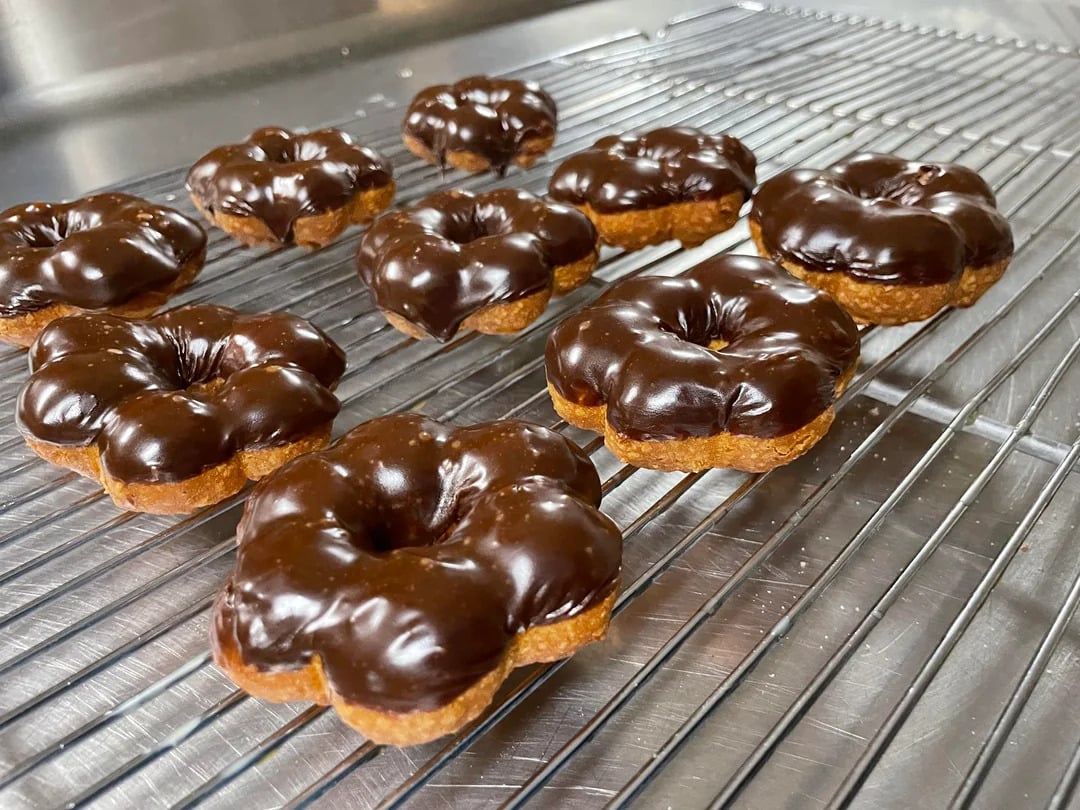 Eight chocolate-glazed flower-shaped donuts are cooling on a metal wire rack over a shiny metal surface in a kitchen setting.