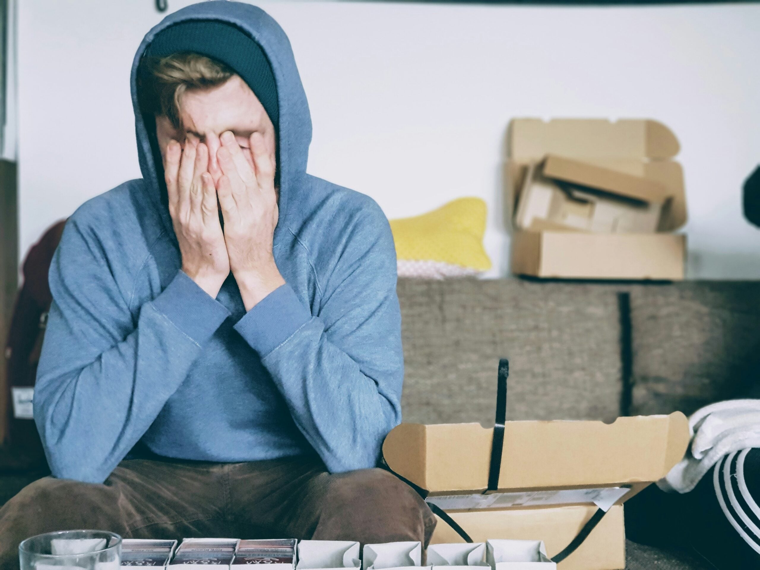A person in a blue hoodie sits on a couch, covering their face with their hands, looking stressed—perhaps overwhelmed by the life in 2026 problems looming ahead. Open cardboard boxes and papers are scattered on the table in front of them.
