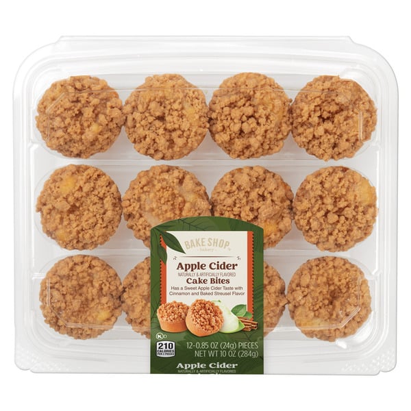 A clear plastic container holds 12 apple cider cake bites with crumb topping. The green label in the center shows the product name and a photo of the cake bites with apple and cinnamon sticks.
