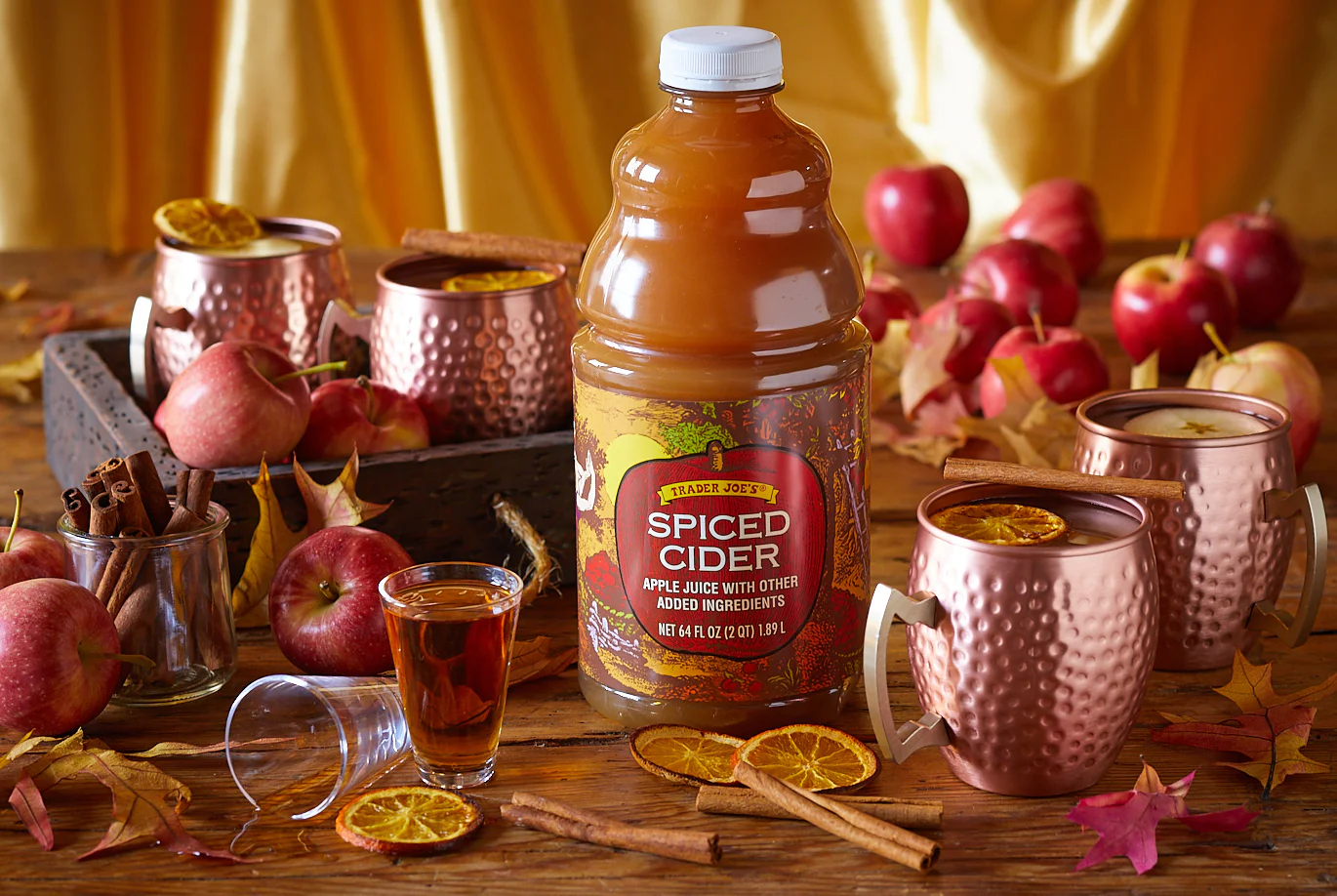 A jug of Trader Joe’s Spiced Cider is surrounded by copper mugs, apples, cinnamon sticks, autumn leaves, and dried orange slices on a rustic wooden table with a yellow cloth in the background.
