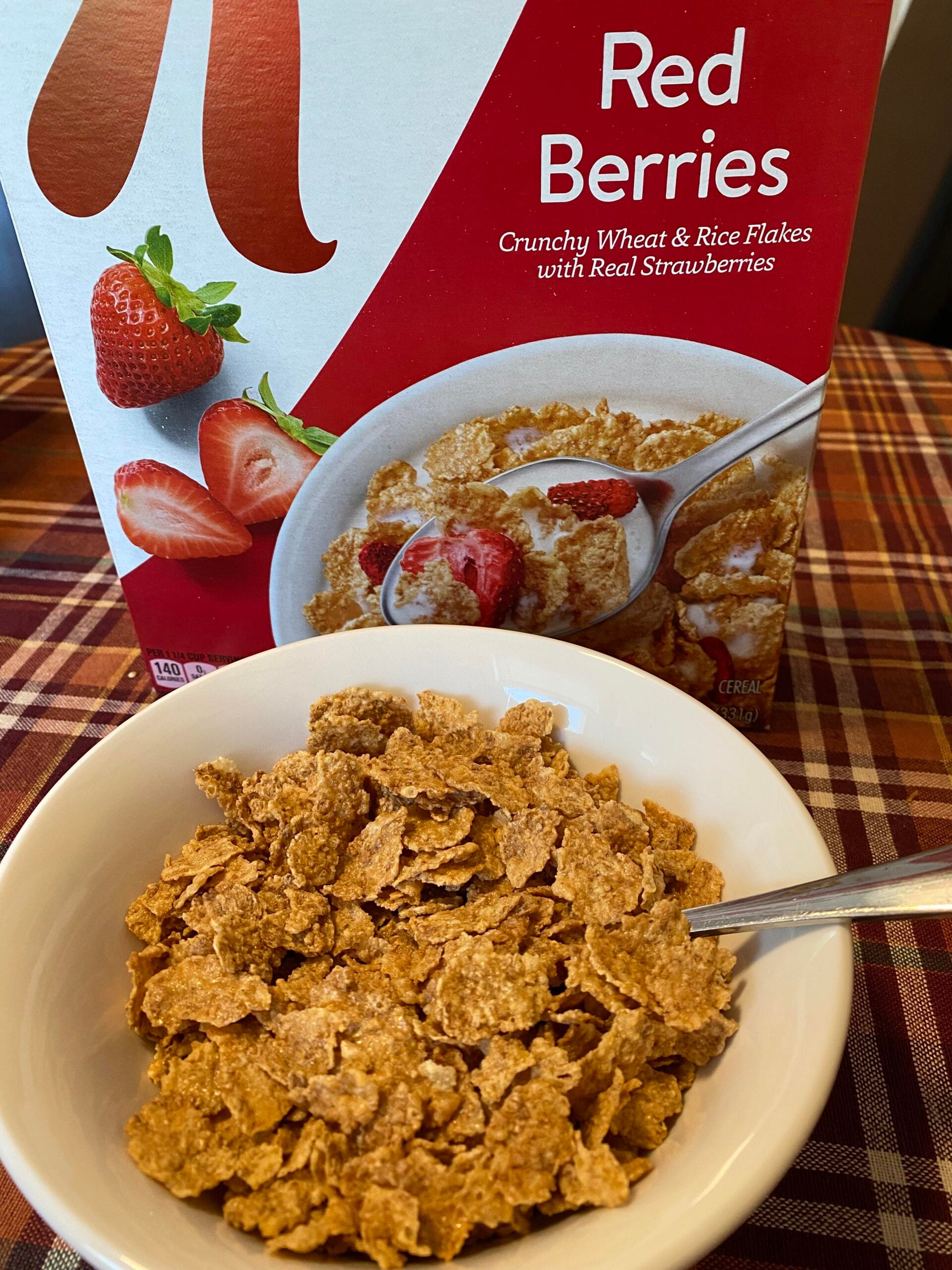 A bowl of wheat and rice flakes cereal sits in front of a Red Berries cereal box on a plaid tablecloth. A spoon rests in the bowl, and the cereal box shows flakes with strawberries.
