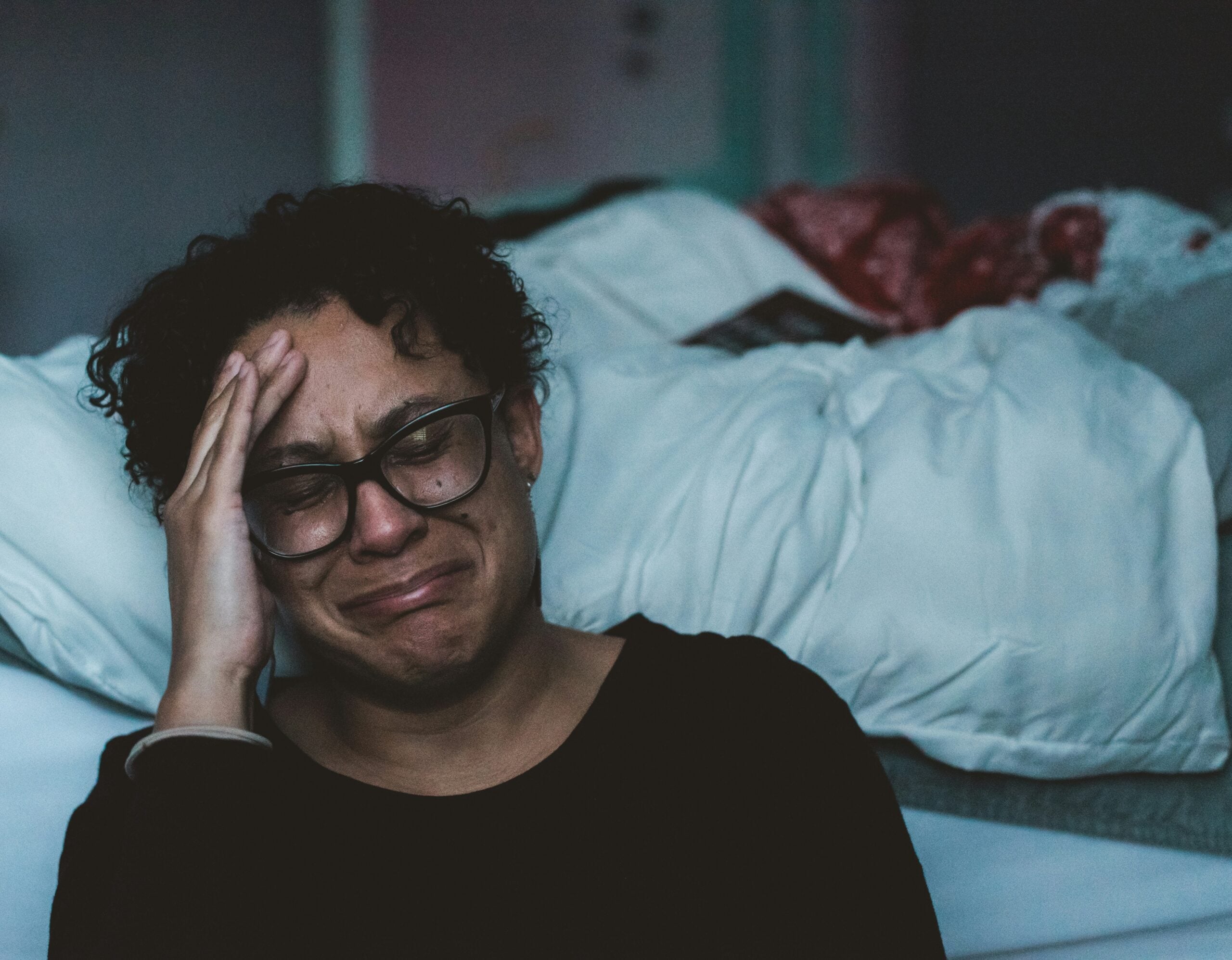 A person wearing glasses sits on the floor by a bed, holding their head in distress and crying. The room is dimly lit, capturing the somber and emotional atmosphere that reflects some of the challenges faced in life in 2026.