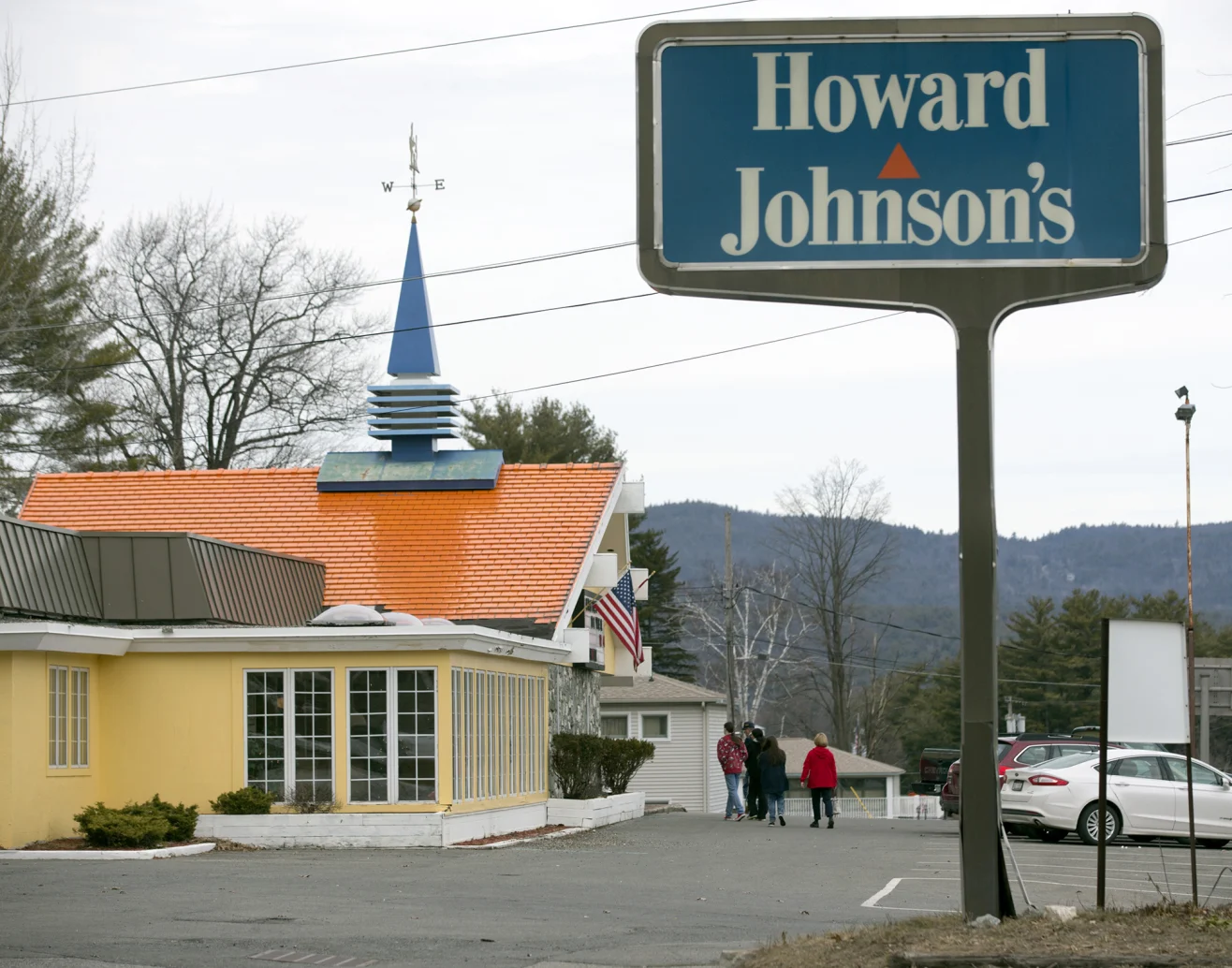 Nostalgic restaurant chains. Howard Johnson’s restaurant with an orange roof and blue spire, a large sign in front, a few people walking near the entrance, and cars in the parking lot. Trees and hills are in the background.