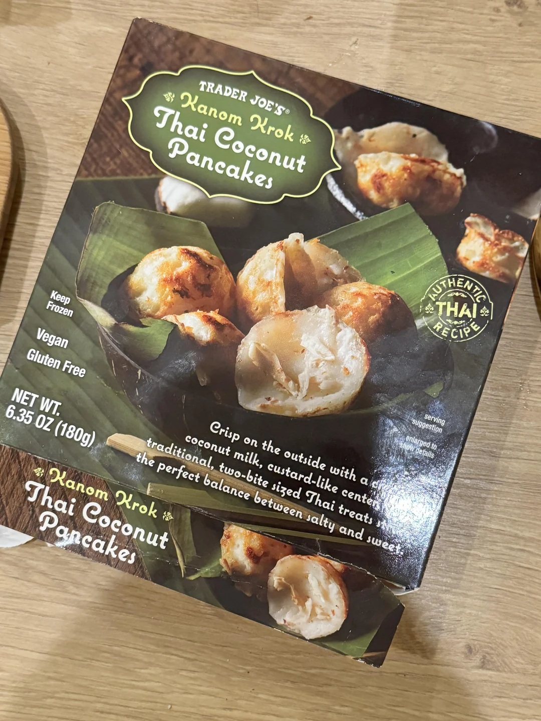 A box of Trader Joe’s Kanom Krok Thai Coconut Pancakes on a wooden surface. The packaging shows images of the round, golden pancakes with creamy filling on green leaves. The box highlights that the product is vegan and gluten-free.