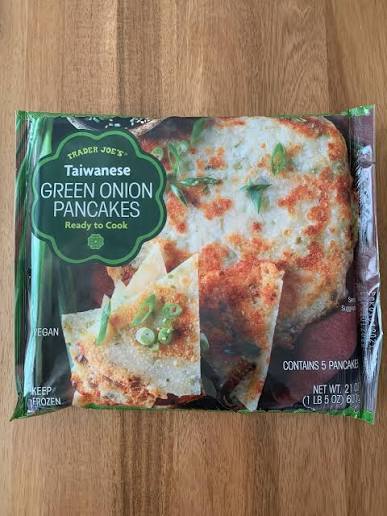 A package of Trader Joe’s Taiwanese Green Onion Pancakes sits on a wooden surface. The packaging shows images of crispy pancakes topped with chopped green onions. The text notes it is vegan, contains 5 pancakes, and is ready to cook.