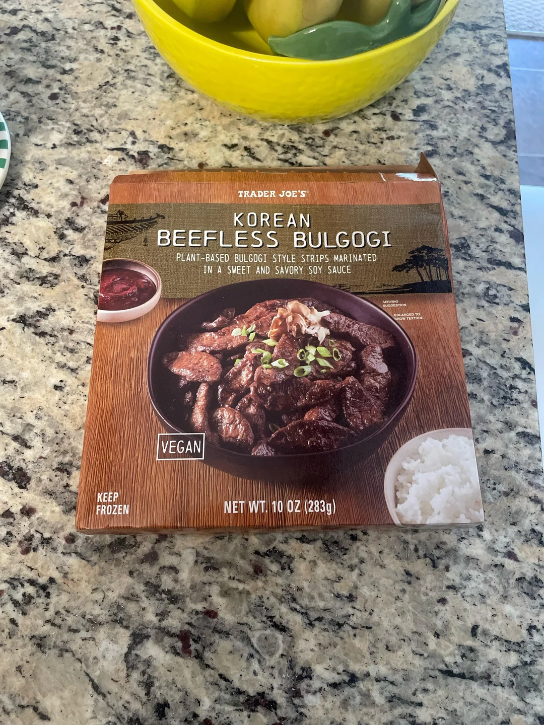 A box of Trader Joe’s Korean Beefless Bulgogi sits on a granite counter. The packaging shows vegan beefless strips with green onions, sauce, and a bowl of white rice. A bowl of lemons is in the background.