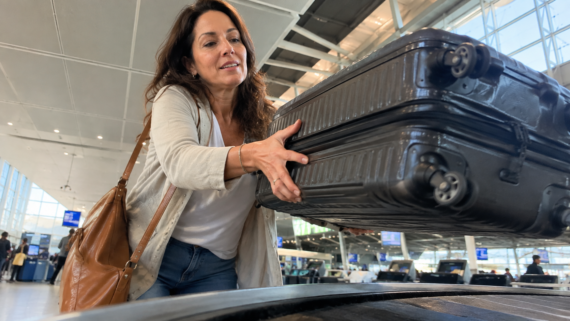 A woman reaches to pick up a black suitcase from a baggage carousel at an airport, ready to begin her vacation. She is wearing a tan jacket and carrying a brown shoulder bag. The terminal is bright and modern with large windows.