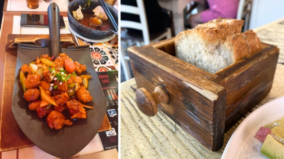 A dish of stir-fried food is served on a mini shovel, showcasing some truly ridiculous food plating, while rustic bread slices are presented in a wooden drawer box on a restaurant table.
