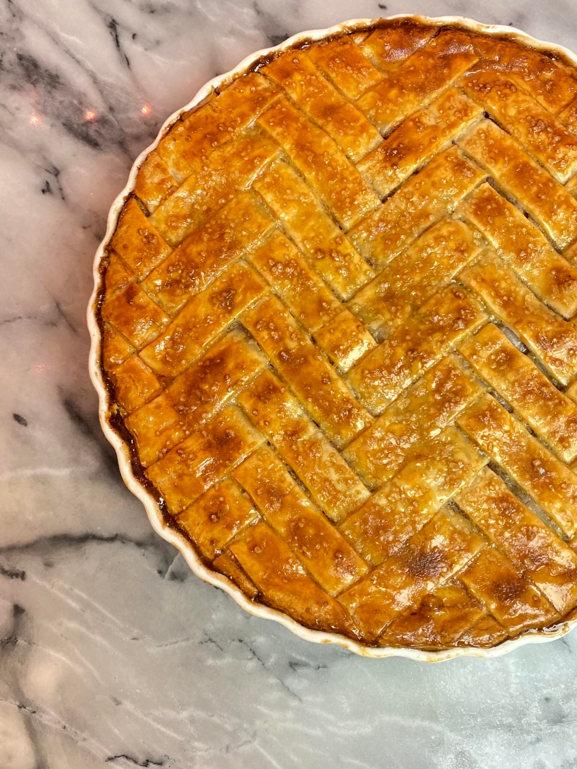 A golden-brown pie with a lattice crust sits in a white dish on a marble surface.