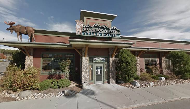A steakhouse with a log cabin design, large front windows, and animal statues—a moose and a deer—on the roof. The sign reads “Bugaboo Creek Steak House.” Shrubs and rocks line the building’s entrance.