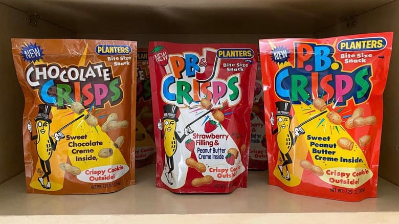 Three Planters snack bags on a shelf: Chocolate Crisps, PB&J Crisps, and PB Crisps. Each bag features Mr. Peanut and highlights crispy cookie snacks with various creamy fillings.