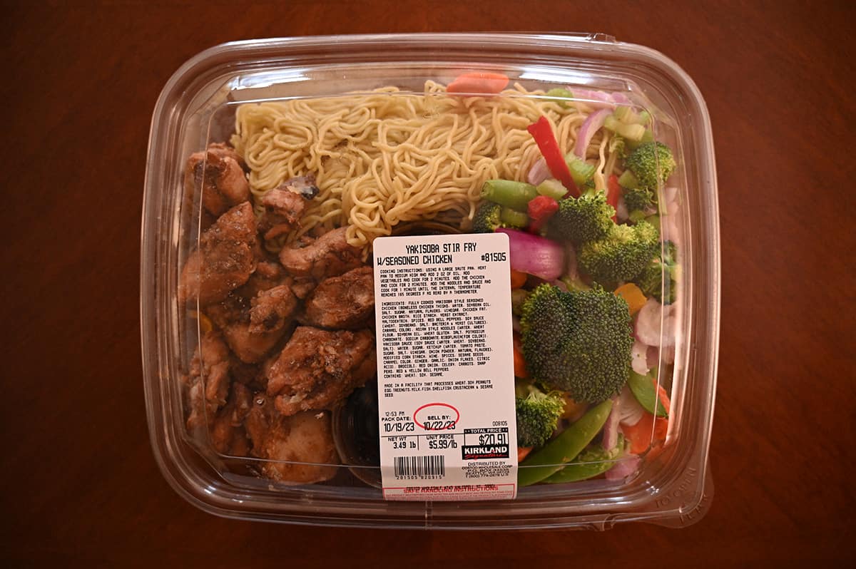 Plastic container with precooked seasoned chicken, noodles, and fresh vegetables like broccoli, carrots, and snap peas. A label with nutrition and product information is on top. The container sits on a wooden surface. Costco foods cheaper than takeout.