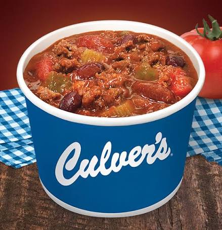 A blue Culver’s cup filled with chili containing beans, ground meat, tomatoes, and celery, sitting on a wooden surface with a blue checkered cloth and a tomato in the background.