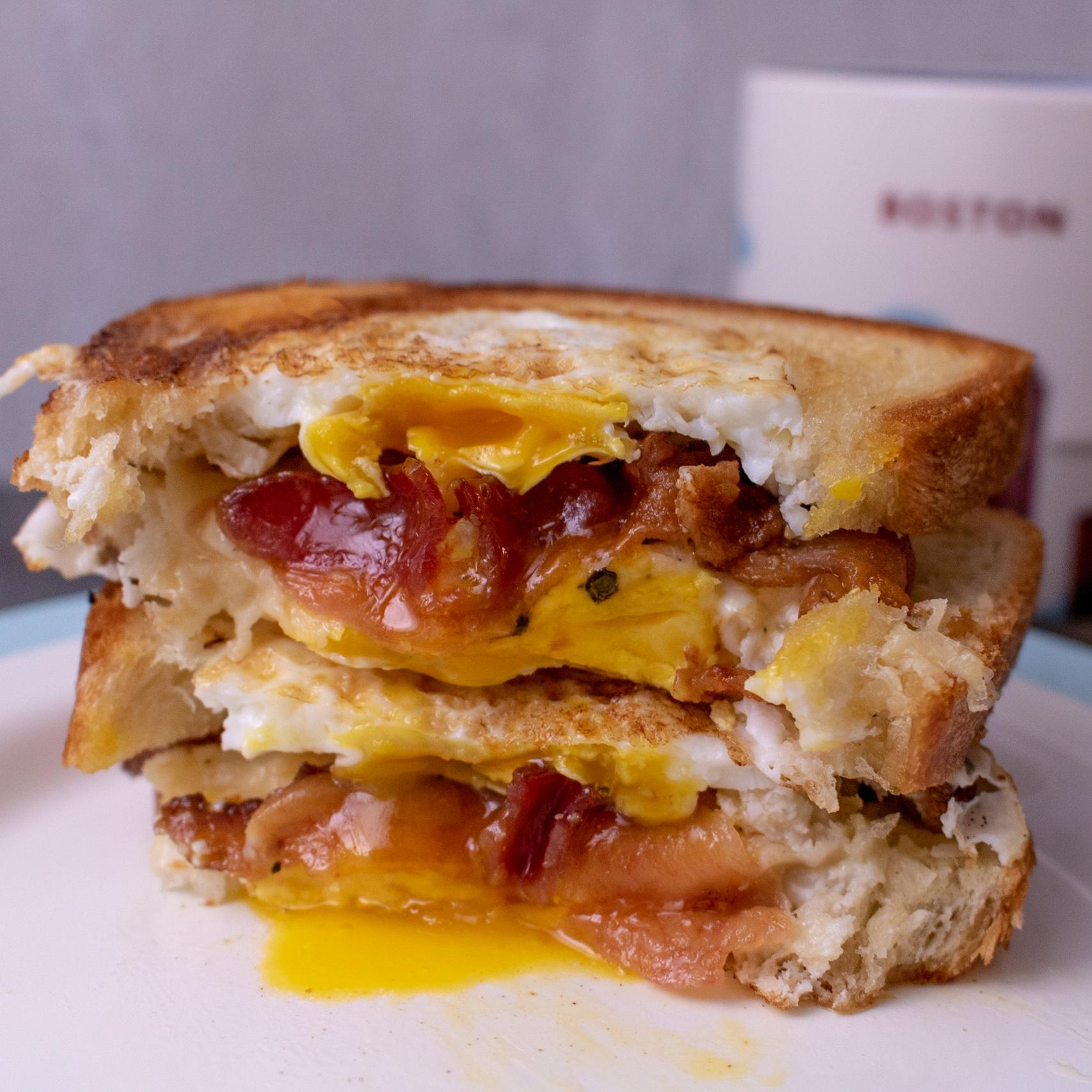 A close-up of a breakfast sandwich with egg, bacon, and tomato on toasted bread, with a bite taken out. The egg yolk is runny and dripping onto the plate. A mug is blurred in the background.